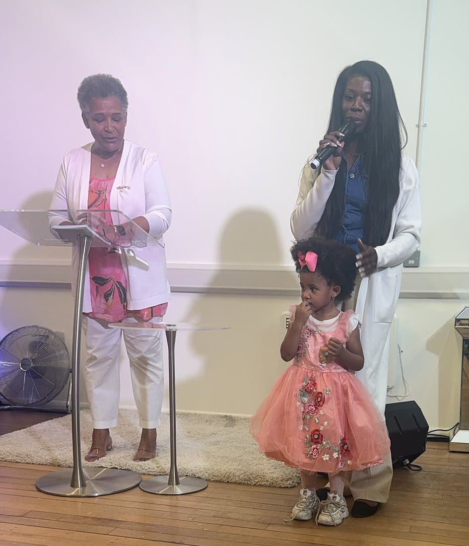 Minister Iris praying at the lectern as a little girl in pink stands with her mother